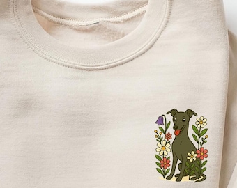 Könnte beinhalten: Ein cremefarbenes Sweatshirt mit einer Grafik eines Hundes, der von bunten Blumen umgeben ist. Der Hund ist dunkelgrün mit roter Zunge, und die Blumen umfassen weiße, gelbe und rote Blüten. Das Sweatshirt hat einen Rundhalsausschnitt und lange Ärmel.