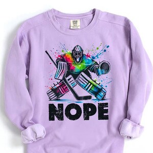 May include: A purple sweatshirt with a colorful graphic of a hockey goalie making a save. The goalie is wearing a helmet and pads. The graphic is surrounded by colorful paint splatters. The text "NOPE" is printed below the graphic.