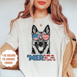 May include: A cream-coloured t-shirt with a German Shepherd dog wearing sunglasses in the style of the American flag. The word "'MERICA" is printed below the dog in red, white, and blue. The shirt also has the text "+10 COLOURS TO CHOOSE FROM!"