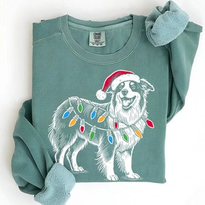Puede incluir: Una sudadera verde con un gráfico blanco de un perro con un gorro de Papá Noel y luces de Navidad. La etiqueta dice "COMFORT COLORS".