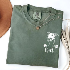 以下が含まれることがあります： 緑色のTシャツに、犬が上の方から顔を出している白いイラストが描かれています。イラストの下には、犬の名前「ボルト」が印刷されています。