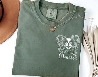 Puede incluir: Una camiseta verde con un gráfico blanco de un perro Papillón asomándose por la parte superior de la camiseta. El texto "Monarch" está impreso debajo del perro.
