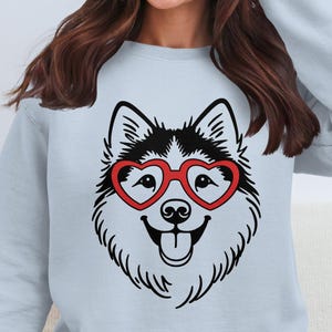 May include: A light blue sweatshirt featuring a black and white illustration of a dog's face wearing red heart-shaped glasses. The dog has a happy expression with its tongue sticking out. The sweatshirt is a casual style.
