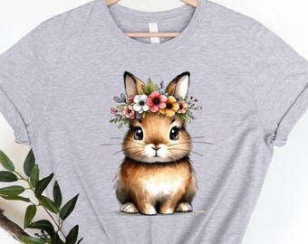 Puede incluir: Una camiseta gris con un lindo conejito de dibujos animados que lleva una corona de flores. El conejito es marrón con pelaje blanco y tiene grandes ojos marrones. Las flores de la corona son rosas, naranjas, amarillas y blancas.