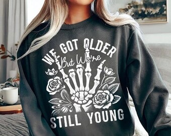 May include: A dark grey sweatshirt with a white graphic print of a skeleton hand giving the middle finger. The text on the sweatshirt reads "We Got Older But We're Still Young".