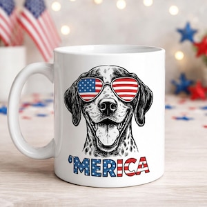 May include: White ceramic mug featuring a black and white illustration of a dog wearing sunglasses with the American flag design. The word "'MERICA" is printed in red, white, and blue. Patriotic theme.