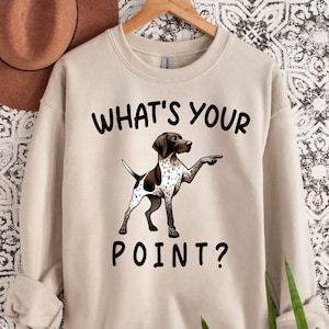 May include: A beige sweatshirt featuring the text "What's your point?" and a cartoon illustration of a brown and white German Shorthaired Pointer dog pointing with its right paw.