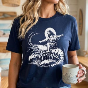 May include: Navy blue t-shirt with a white graphic of a figure with a halo, wearing sunglasses, surfing on a shrimp. The design includes stylized waves. The person in the image is holding a mug with a wave design.