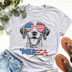May include: A light gray t-shirt featuring a smiling dog wearing sunglasses with an American flag design. The word "'MERICA" is printed below the dog in red, white, and blue. The shirt is paired with blue jeans and brown sandals.