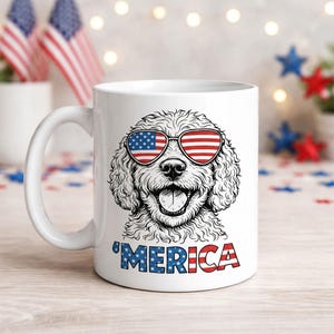Puede incluir: Taza de cerámica blanca con una ilustración en blanco y negro de un perro con gafas de sol con el diseño de la bandera estadounidense. La palabra "'MERICA" está impresa en rojo, blanco y azul debajo.
