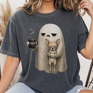 Może przedstawiać: Ciemnoszary t-shirt z krótkim rękawem z grafiką przedstawiającą białego ducha o zrzędliwym wyrazie twarzy, trzymającego brązowy kubek z napisem MEH oraz małego, płowego psa rasy chihuahua. Duch ma ciemne oczy i opadające kąciki ust.