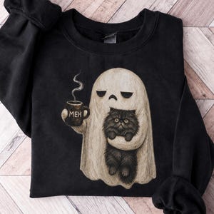 Op de afbeelding: Een zwart sweatshirt met ronde hals voorzien van een afbeelding van een beige geest die een donkere mok met de tekst MEH vasthoudt en een pluizige zwarte kat draagt. De geest heeft een neutrale uitdrukking en er stijgt stoom uit de mok.