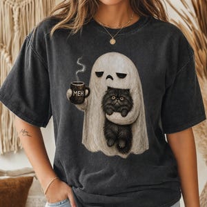 Könnte beinhalten: Ein anthrazitfarbenes Kurzarm-T-Shirt mit einer Grafik eines weißen Geistes, der eine dampfende Tasse mit der Aufschrift MEH hält und eine flauschige schwarze Katze trägt. Der Geist hat einen neutralen Ausdruck und die Katze hat große, gelbe Augen.