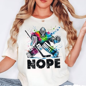 May include: A white t-shirt featuring a colourful graphic of a hockey goalie in action. The goalie is wearing a blue and green jersey with a white helmet. The graphic is surrounded by colourful paint splatters. The text "NOPE" is printed in black below the graphic. The shirt is available in ivory.