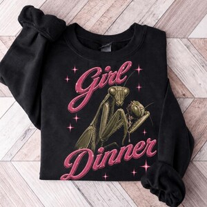 Może przedstawiać: Czarna bluza z długim rękawem z grafiką przedstawiającą jedzącą modliszkę. Napis "Girl Dinner" jest wydrukowany różową czcionką skryptową nad i pod owadem. Wokół centralnego wzoru rozmieszczone są małe, różowe, czteroramienne gwiazdki.