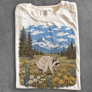 Pode incluir: Uma t-shirt de cor creme com uma impressão gráfica de um guaxinim a caminhar por um campo de flores silvestres amarelas e brancas. Ao fundo, há pinheiros altos e montanhas com neve sob um céu azul com nuvens.