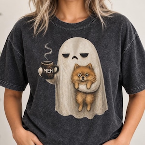 Może przedstawiać: Ciemnoszary t-shirt z efektem sprania, ozdobiony grafiką przedstawiającą białego ducha z znudzonym wyrazem twarzy, trzymającego brązowy kubek z napisem MEH i niosącego puszystego brązowego szpica miniaturowego. Koszulka ma krótki rękaw i okrągły dekolt.