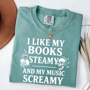 May include: A teal Comfort Colors t-shirt with the text "I LIKE MY BOOKS STEAMY AND MY MUSIC SCREAMY" in white. The shirt features graphic elements of a book, heart, and skull with headphones. A silver necklace with a pendant is draped on the shirt.