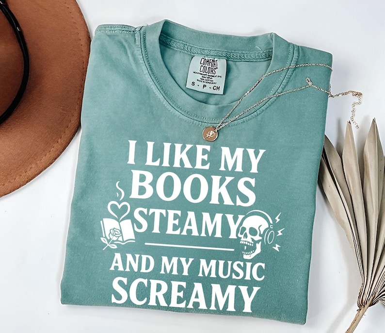 May include: A teal Comfort Colors t-shirt with the text "I LIKE MY BOOKS STEAMY AND MY MUSIC SCREAMY" in white. The shirt features graphic elements of a book, heart, and skull with headphones. A silver necklace with a pendant is draped on the shirt.