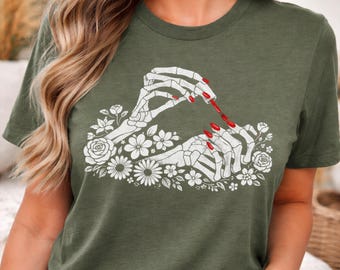May include: Olive green t-shirt featuring a white graphic of skeleton hands applying red nail polish. The hands are surrounded by white floral elements, including roses and daisies. The design is centered on the shirt.