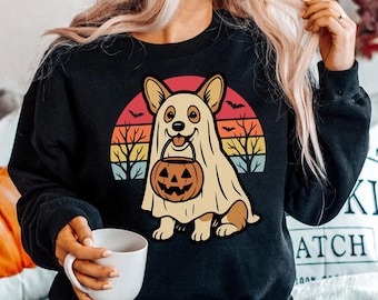 May include: Black sweatshirt featuring a Halloween-themed graphic. The design includes a corgi dressed as a ghost, holding a jack-o'-lantern bucket. A colorful sunset, bats, and bare trees complete the festive illustration. A white mug is held in the hand.