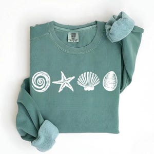 May include: A teal green sweatshirt with a white graphic of a seashell, starfish, and spiral shell. The sweatshirt has a crew neck and long sleeves.