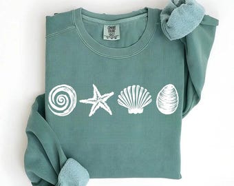Könnte beinhalten: Ein tealgrüner Pullover mit einer weißen Grafik eines Muschel, eines Seesterns und einer spiralförmigen Muschel. Der Pullover hat einen Rundhalsausschnitt und lange Ärmel.