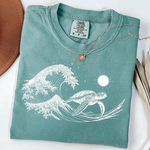 Op de afbeelding: Een tealblauw t-shirt met een witte afbeelding van een zeeschildpad die door een golf zwemt. Het label staat "COMFORT COLORS".