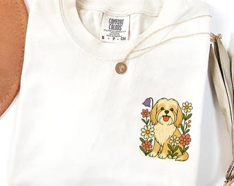 May include: Cream-coloured t-shirt featuring an embroidered design of a light brown dog amidst colourful flowers. The shirt has a crew neck and short sleeves. A gold necklace with a pendant is draped across the shirt.