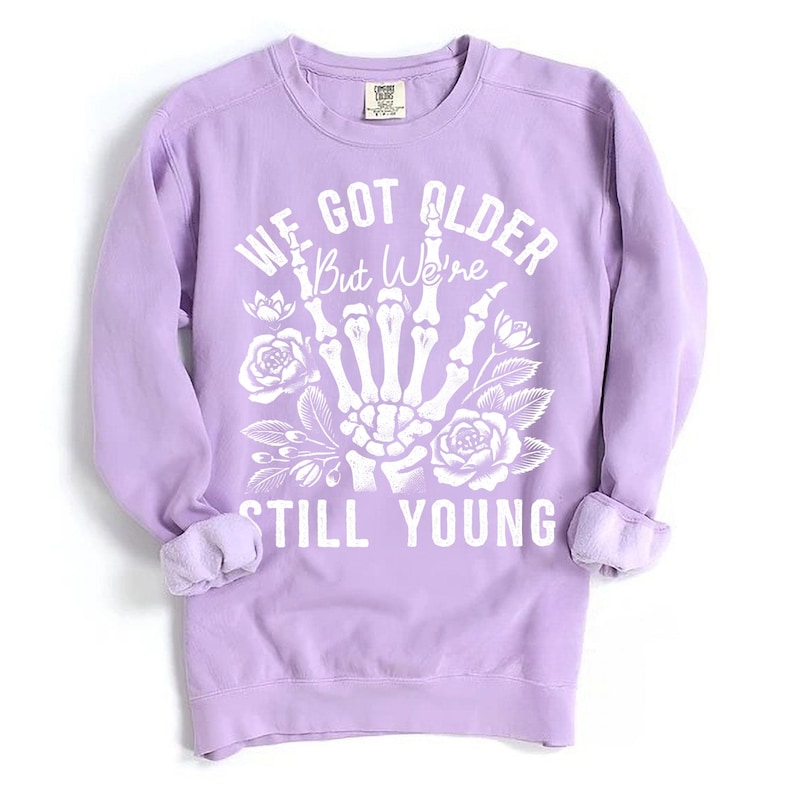 May include: A lavender sweatshirt with a white graphic print of a skeleton hand giving the middle finger with flowers and the text "We got older but we're still young".