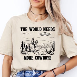 May include: A beige t-shirt with a black graphic print of a cowboy riding a horse in front of a desert landscape. The text "The World Needs More Cowboys" is printed below the graphic. A UFO is flying above the cowboy.