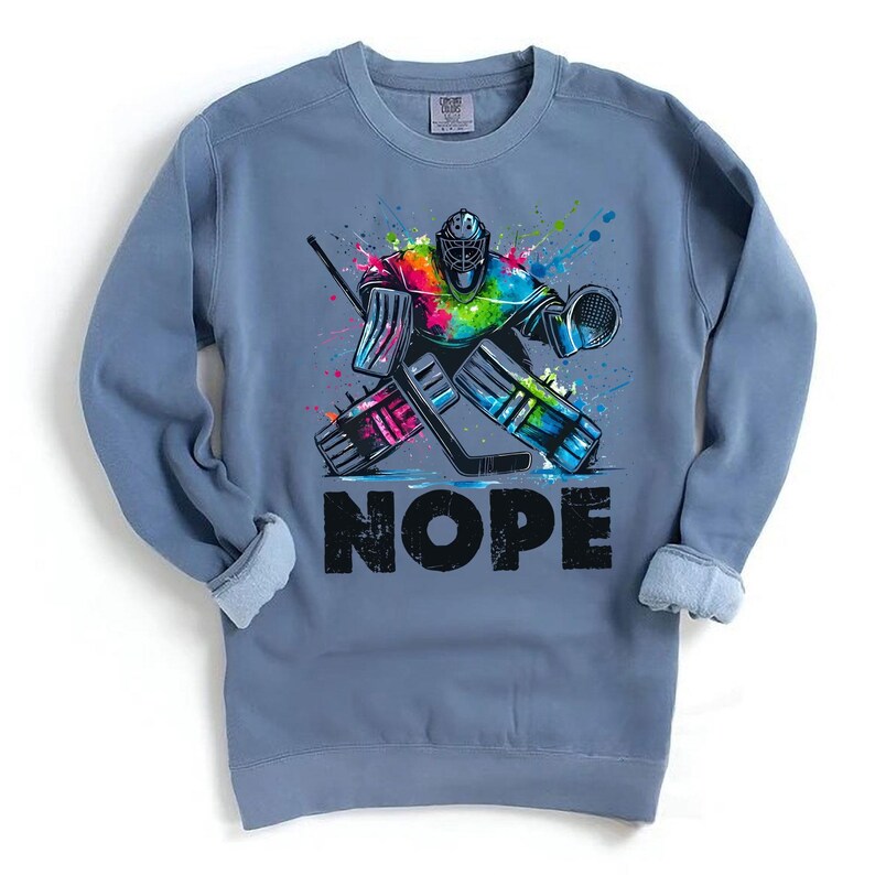 May include: A blue sweatshirt with a colorful graphic of a hockey goalie making a save. The goalie is wearing a helmet and pads. The graphic is surrounded by colorful paint splatters. The text "NOPE" is printed below the graphic.