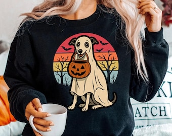 Puede incluir: Sudadera negra con diseño de Halloween. Muestra un perro disfrazado de fantasma sosteniendo un cubo de calabaza, sobre un fondo de puesta de sol con murciélagos y árboles. La persona sostiene una taza blanca. Adecuado para la temporada de otoño.