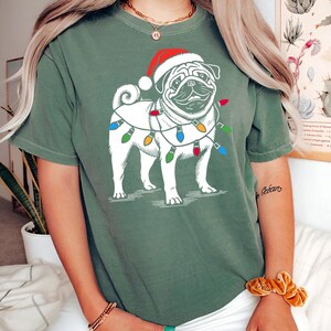 May include: Green short-sleeved t-shirt with a white pug wearing a Santa hat and Christmas lights around its neck.