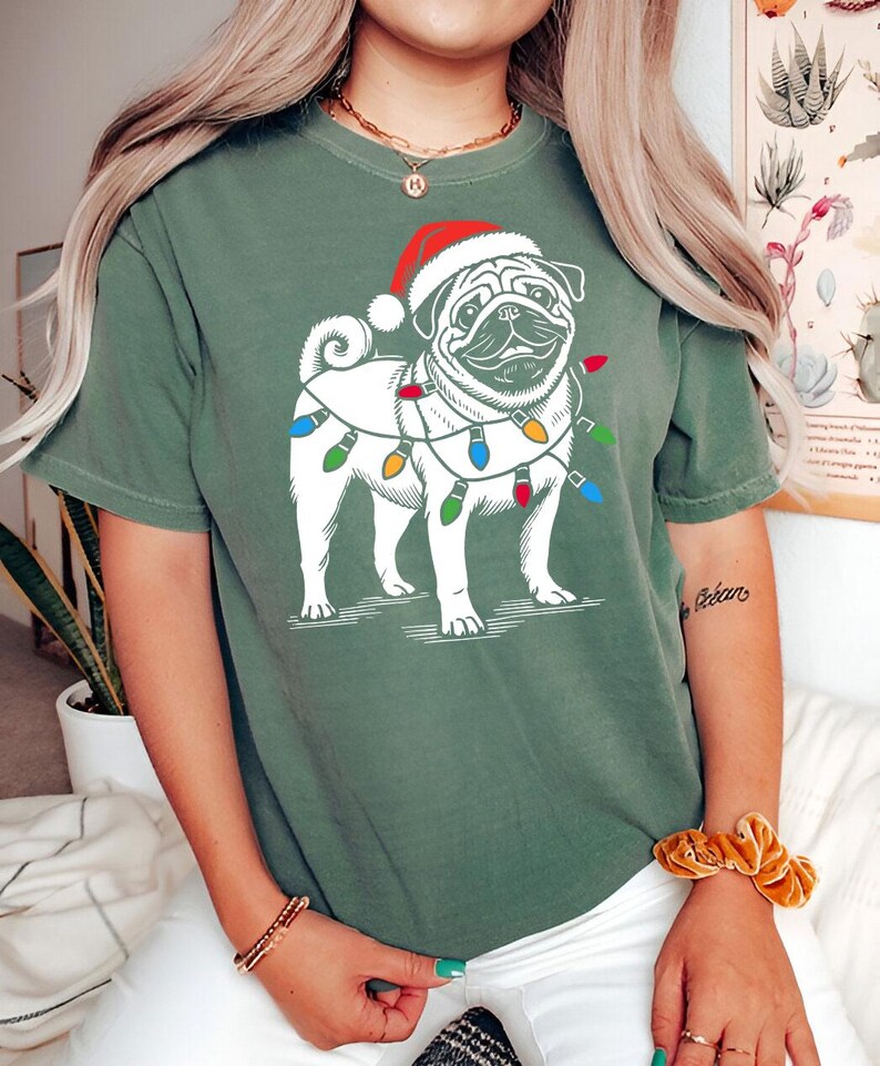 May include: Green short-sleeved t-shirt with a white pug wearing a Santa hat and Christmas lights around its neck.