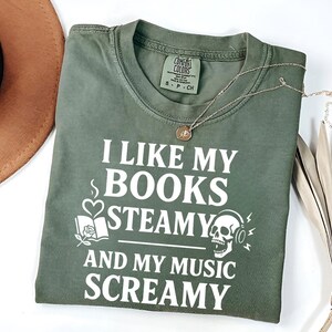 May include: Olive green t-shirt with the text "I LIKE MY BOOKS STEAMY AND MY MUSIC SCREAMY" in white. The shirt has a small tag at the neckline and a silver necklace. A brown hat and dried plant are in the background.