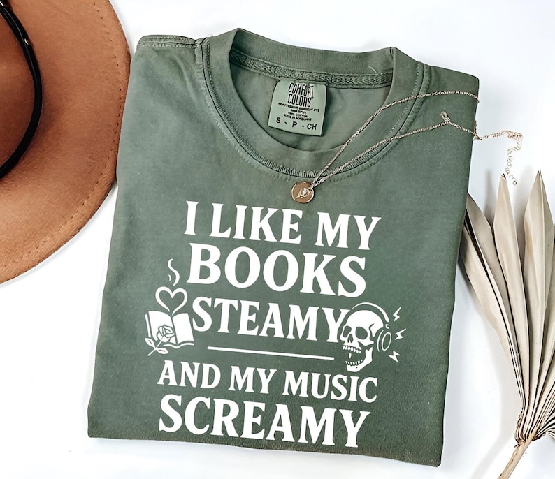 May include: Olive green t-shirt with the text "I LIKE MY BOOKS STEAMY AND MY MUSIC SCREAMY" in white. The shirt has a small tag at the neckline and a silver necklace. A brown hat and dried plant are in the background.