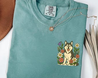 May include: A teal Comfort Colors t-shirt with a graphic of a smiling husky dog surrounded by flowers. The shirt has a small tag with the brand name and size. A silver necklace with a pendant is draped across the shirt.