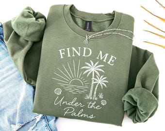 Może przedstawiać: Zielona bluza w kolorze oliwkowym z białym napisem "FIND ME Under the Palms". Projekt zawiera słońce, palmy i muszle. Srebrny naszyjnik jest zarzucony na bluzę, która jest połączona z niebieskimi dżinsami.