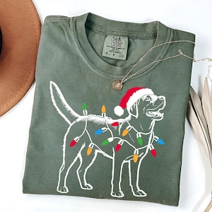 Puede incluir: Una camiseta verde con un contorno blanco de un perro con un gorro de Papá Noel y luces de Navidad. El texto "COMFORT COLORS" está en una etiqueta adjunta a la camiseta.
