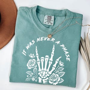 May include: A teal blue t-shirt with a white graphic of a skeleton hand making the rock and roll sign with flowers around it. The text "It was never a phase" is written around the hand.