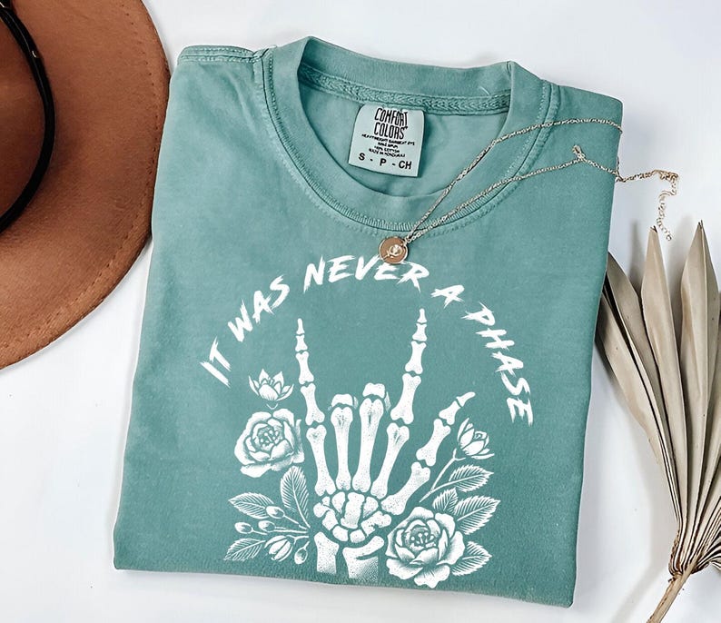May include: A teal blue t-shirt with a white graphic of a skeleton hand making the rock and roll sign with flowers around it. The text "It was never a phase" is written around the hand.