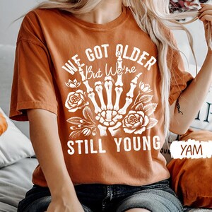 Pode incluir: Uma camiseta laranja queimada com uma m&atilde;o de esqueleto branca fazendo o dedo do meio. O texto na camiseta diz "We got older but we're still young".