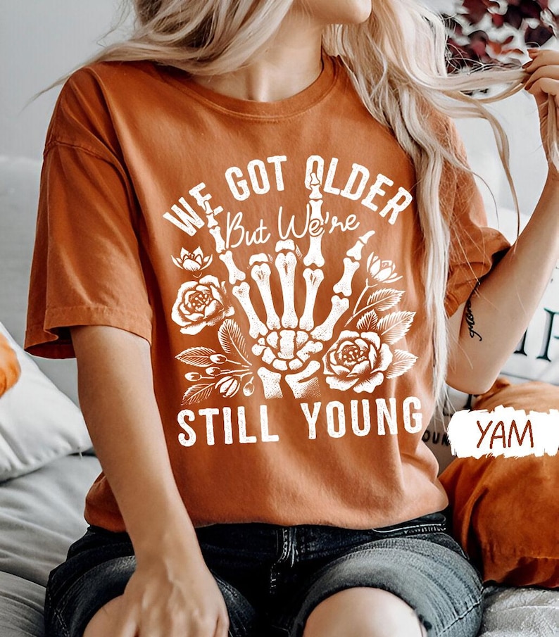 Pode incluir: Uma camiseta laranja queimada com uma m&atilde;o de esqueleto branca fazendo o dedo do meio. O texto na camiseta diz "We got older but we're still young".