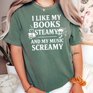 May include: Olive green t-shirt with the text "I LIKE MY BOOKS STEAMY AND MY MUSIC SCREAMY" in white. The graphic includes a book, heart, skull, and headphones. The shirt has short sleeves and a crew neck.