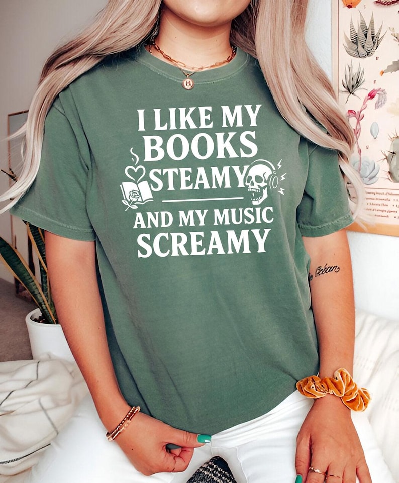 May include: Olive green t-shirt with the text "I LIKE MY BOOKS STEAMY AND MY MUSIC SCREAMY" in white. The graphic includes a book, heart, skull, and headphones. The shirt has short sleeves and a crew neck.