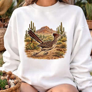 May include: White sweatshirt with a desert-themed graphic. The design shows a roadrunner bird with brown and white plumage, set against a backdrop of cacti and a mountain range. The bird is in motion, with a red crest.