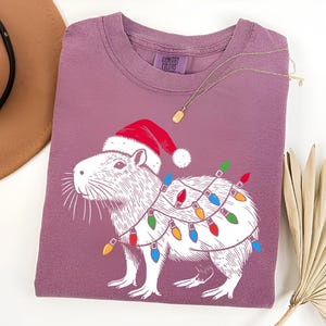 May include: A purple t-shirt with a white illustration of a capybara wearing a Santa hat and Christmas lights. The text "COMFORT COLORS" is visible on the shirt.