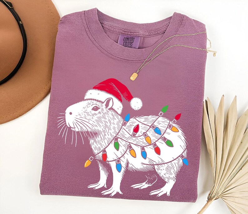 May include: A purple t-shirt with a white illustration of a capybara wearing a Santa hat and Christmas lights. The text "COMFORT COLORS" is visible on the shirt.