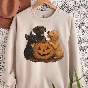 May include: Beige sweatshirt featuring a Halloween-themed graphic. The design includes a carved pumpkin with a smiling face, surrounded by a black French bulldog, a brown Labrador, and a golden retriever. The sweatshirt is hanging on a wooden hanger.